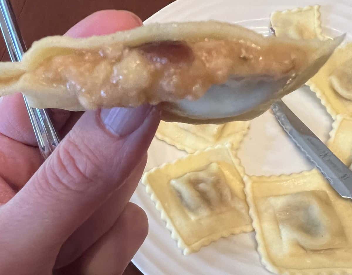 Image of a hand holding one ravioli piece close to the camera that's cut in half so you can see the filling. 