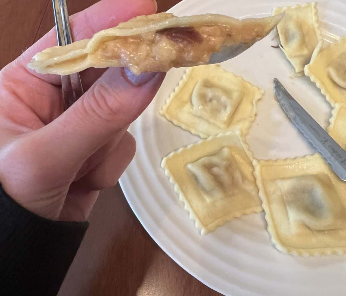 Image of a hand holding one ravioli that's been cut in half hovering over a plate of ravioli so you can see the filling. 