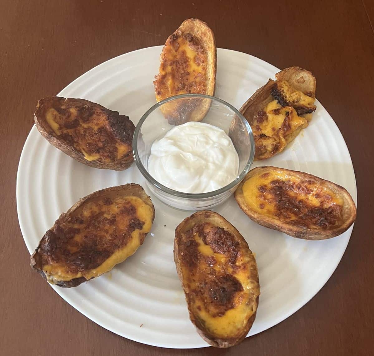 Image of six potato skins cooked and served on a white plate with sour cream in the middle.