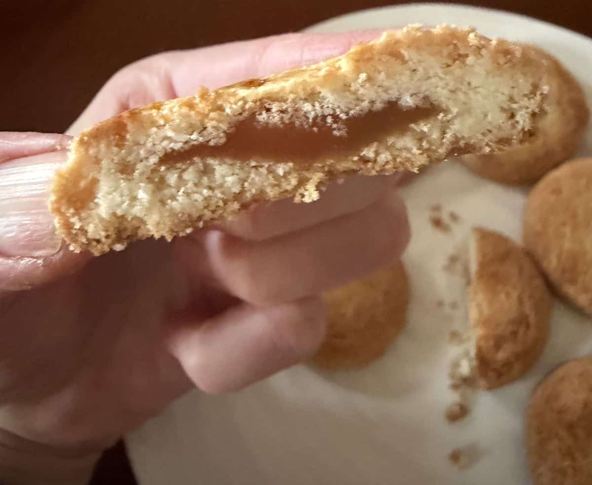 Image of a hand holding one butter cookie close to the camera with a bite taken out of it so you can see the caramel filling.