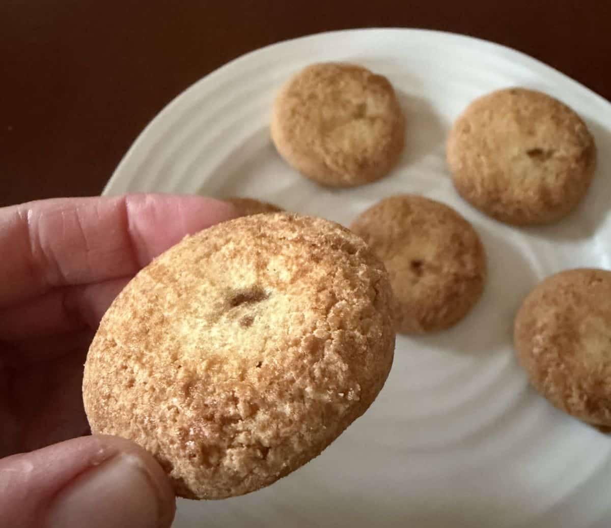 Closeup image of a hand holding one butter cookie close to the camera with a plate of butter cookies in the background. 