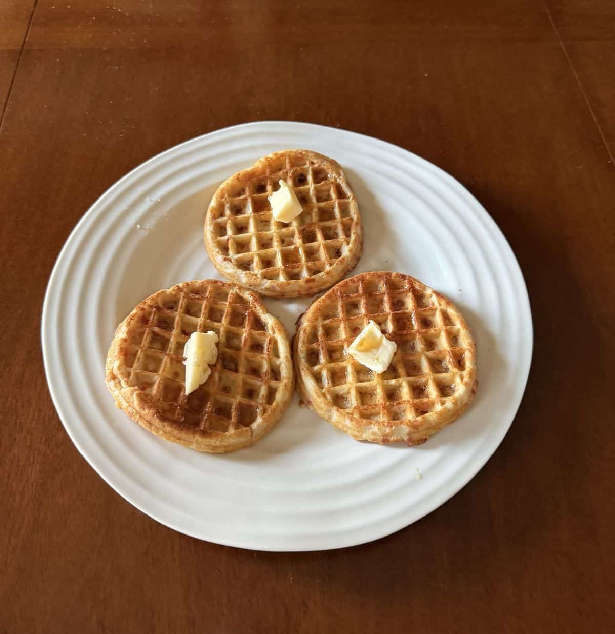 Image of three waffles toasted and served on a white plate with butter and syrup on them.