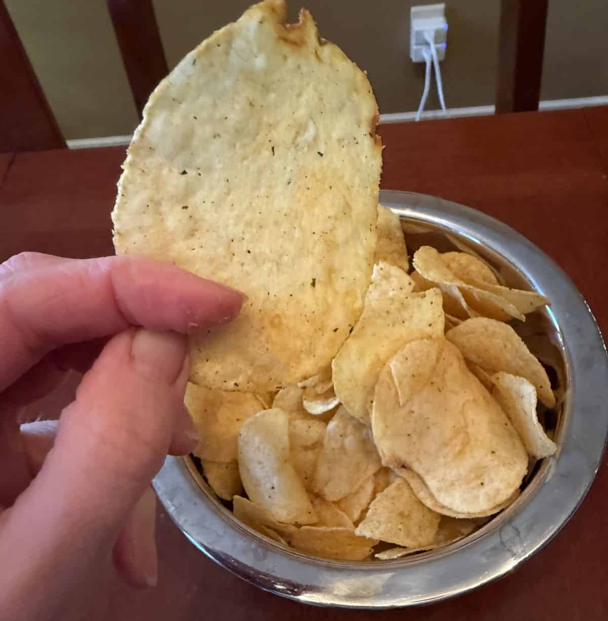 Image of a hand holding a chip close to the camera with a bowl of chips in the background of the photo. 