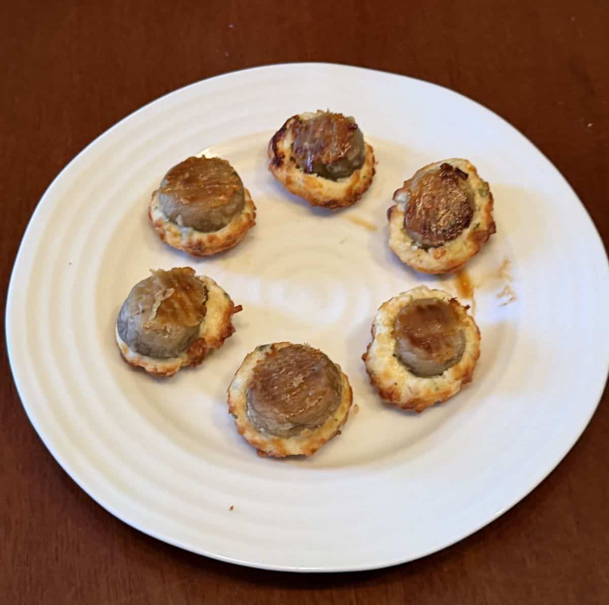 Image of six stuffed mushrooms cooked and served on a white plate upside down so you can see the mushroom bottoms.