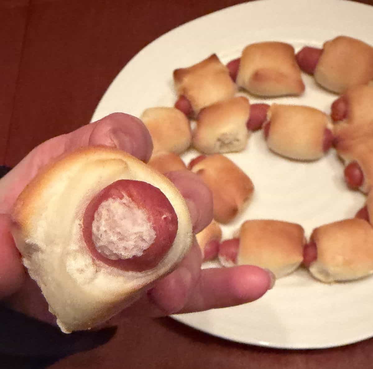 Image of a hand holding one mini sausage in soft baked bread close to the camera with a plate of mini sausages in the background.