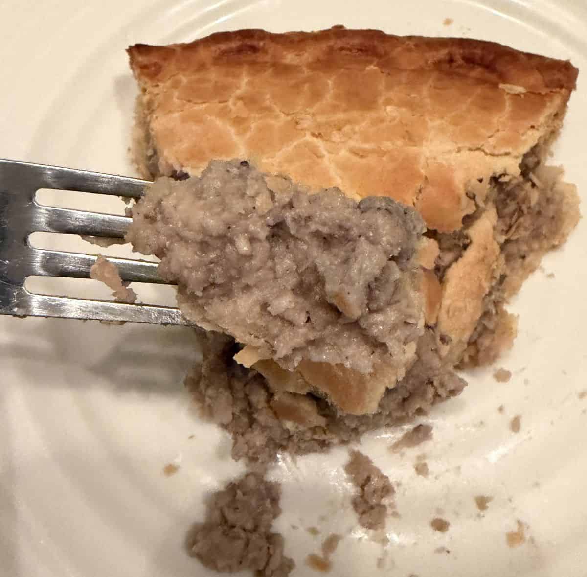 Image of a fork with a bite of meat on it hovering over a slice of meat pie on a white plate.