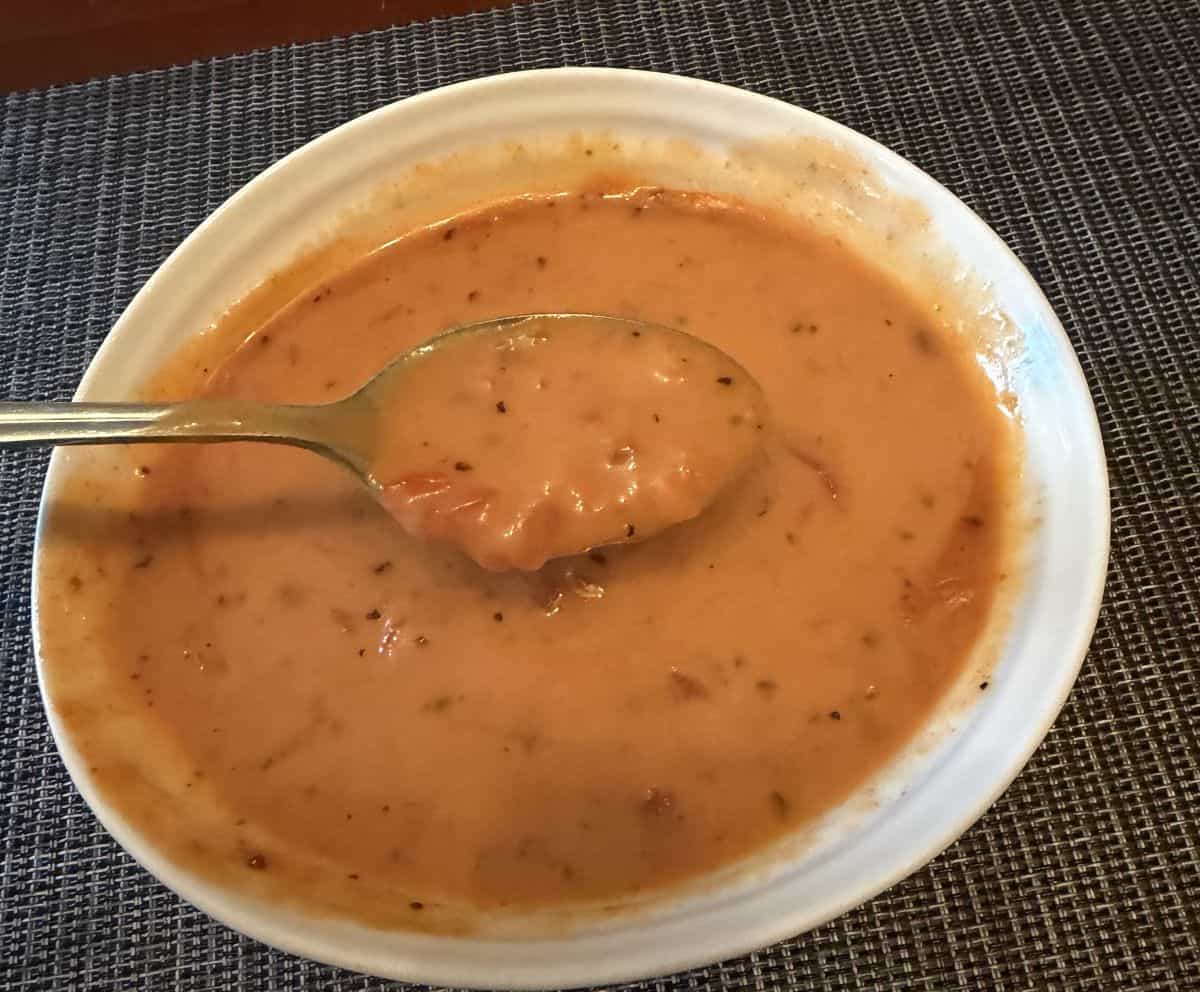 Top down image of a spoon with soup on it hovering over a bowl of soup. There are lots of chunks of red pepper in the soup.