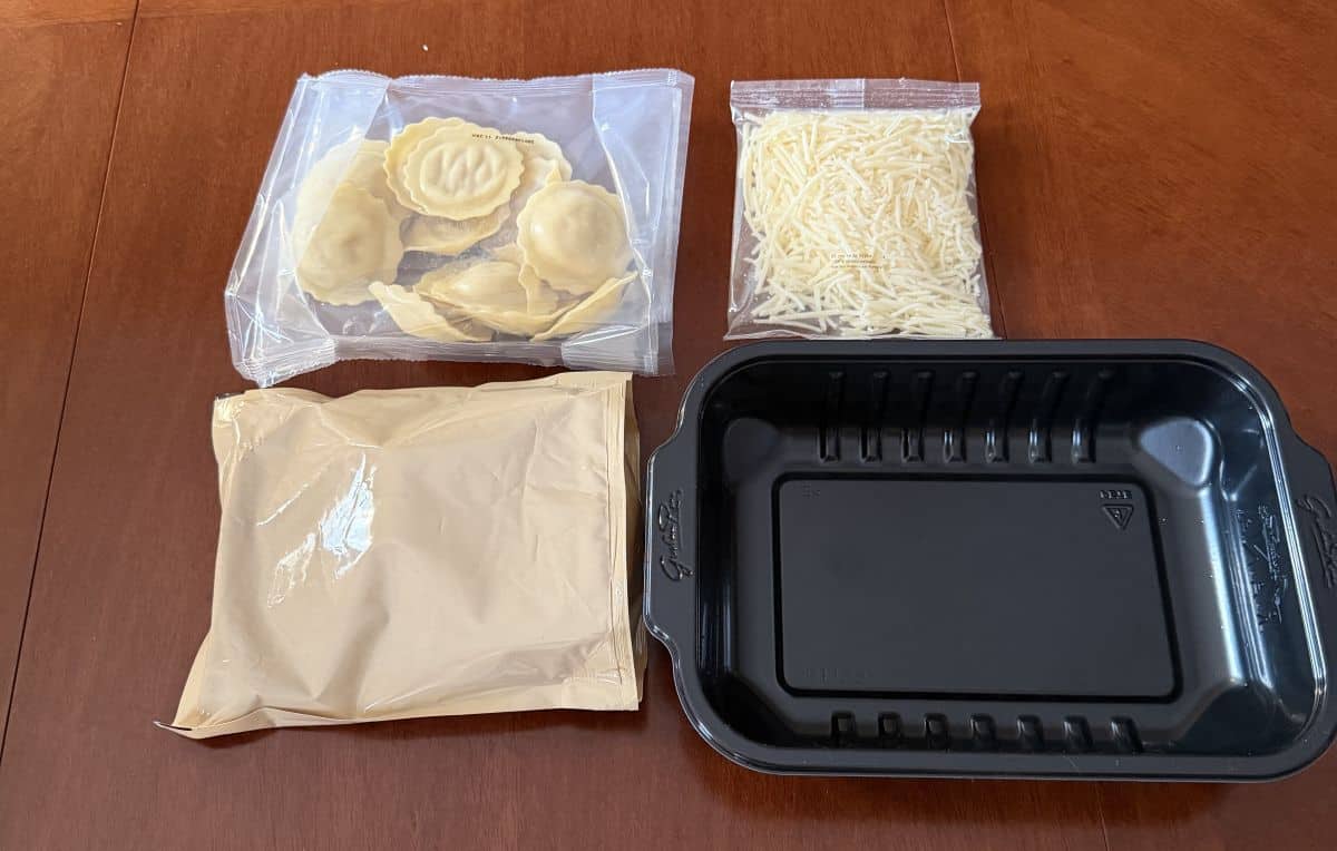 Image of the contents of the pasta kit on a table, cheese, sauce, ravioli and a plastic tray.