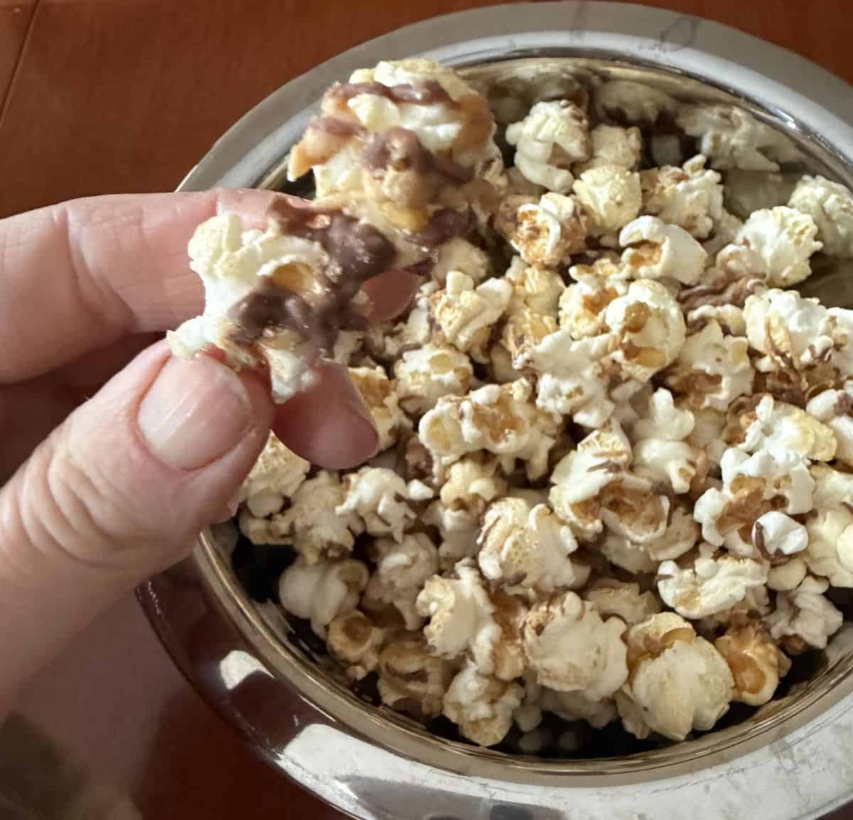Image of a hand holding one piece of Reese's popcorn close to the camera with a bowl of popcorn in the background.