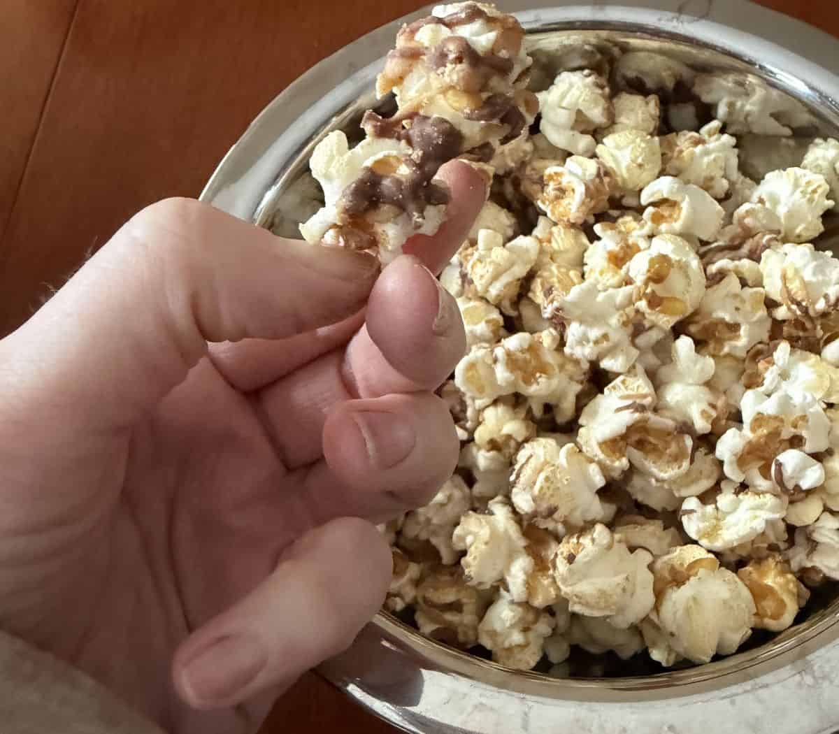 Image of a hand holding one piece of Reese's popcorn very close to the camera with a bowl of popcorn in the background.
