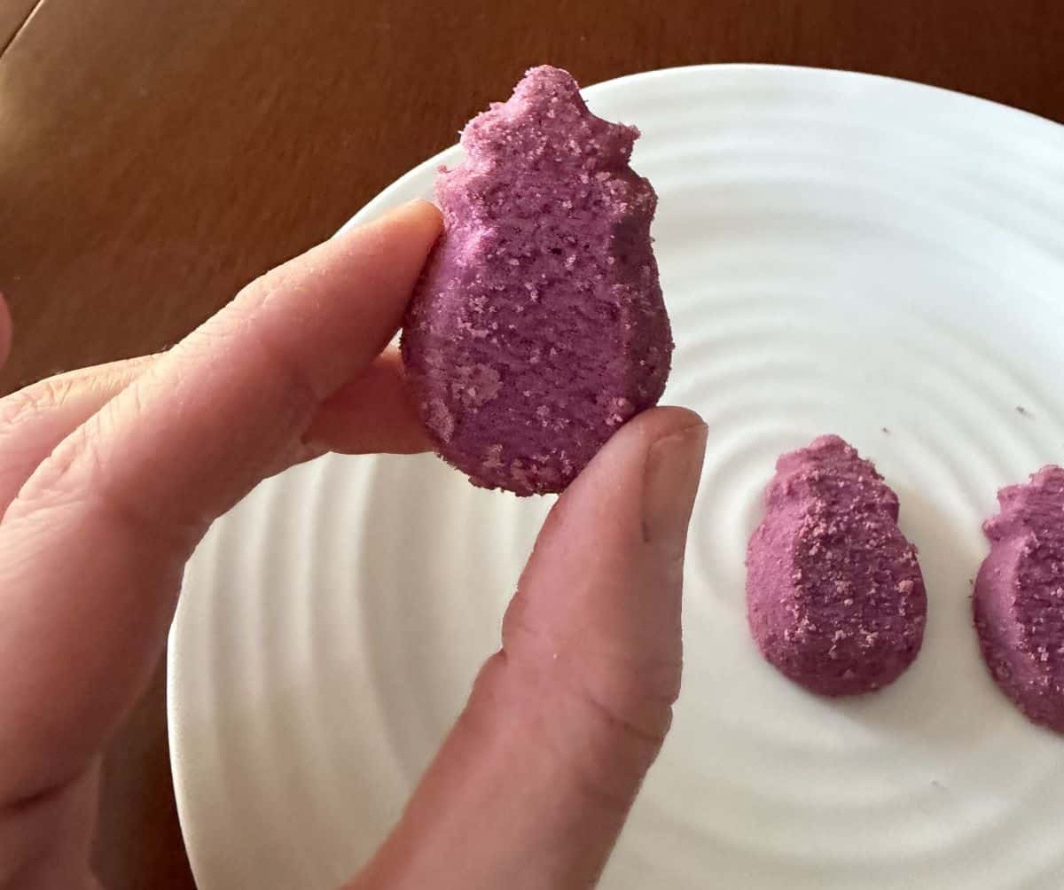Image of a hand holding one ube shortbread bite close to the camera with a plate of ube shortbread in the background.