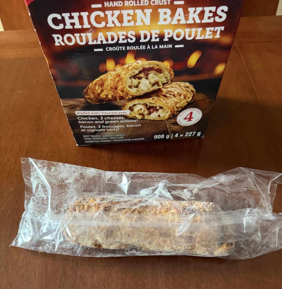 Image of one chicken bake unopened and individually wrapped in plastic sitting in front of the box of chicken bakes on a table.