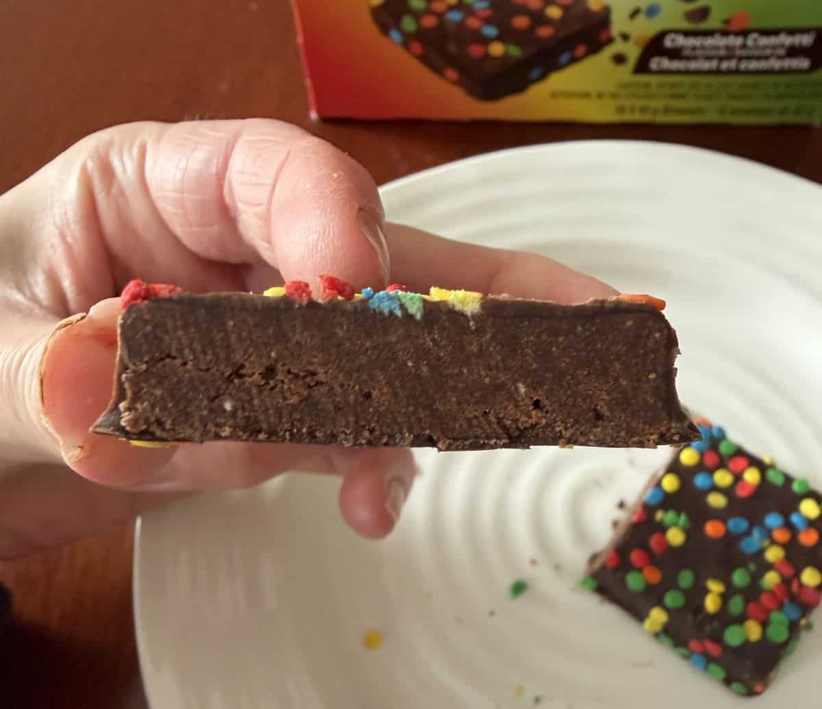 Image of a hand holding one brownie close to the camera with a bite taken out of it so you can see what the inside looks like.