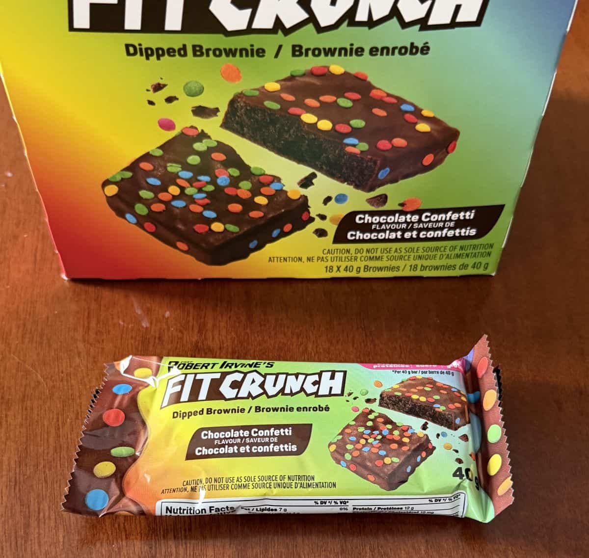 Image of one individually packaged unopened protein brownie sitting on the table in front of the box.