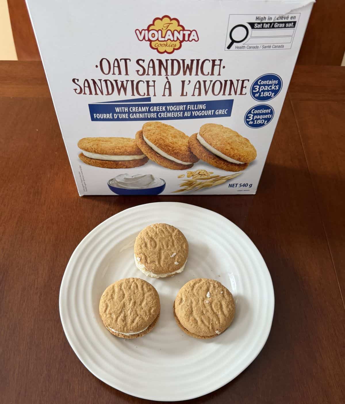 High up top down image of three oat cookies served on a white plate in front of the box of the oat sandwich cookies.
