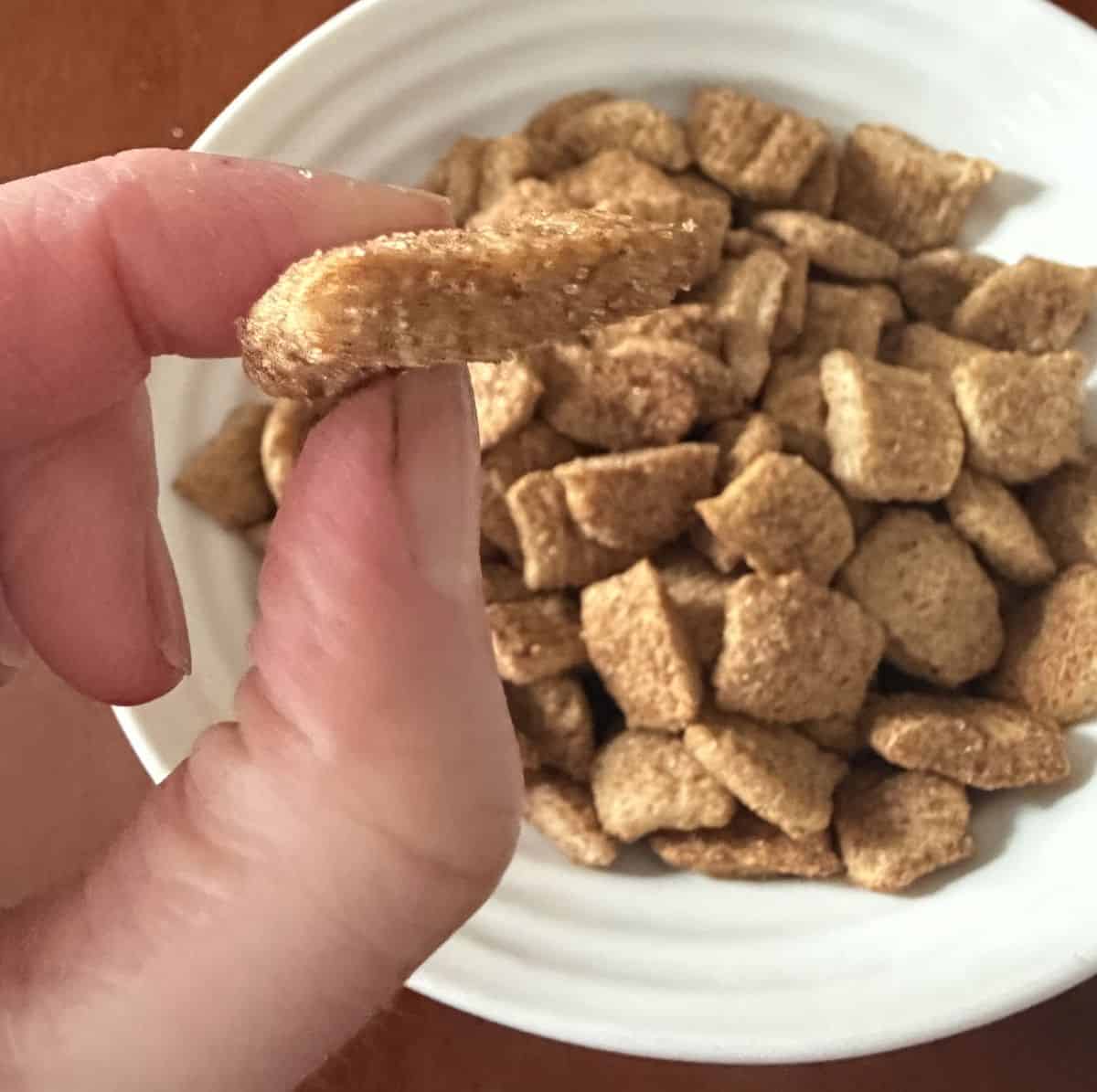 Image of a hand holding up one piece of cereal hovering over a bowl of the cereal.