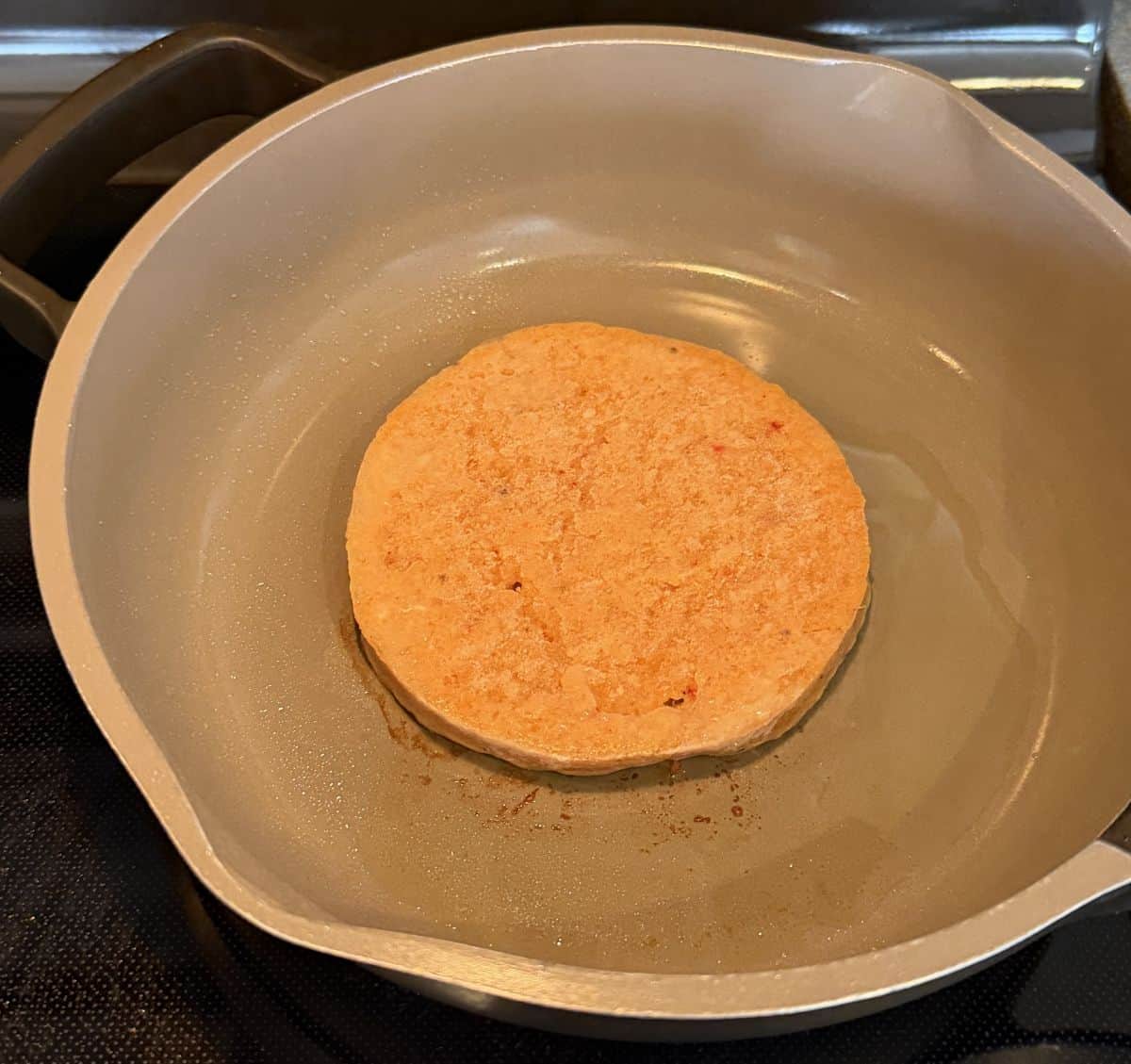 Image of one frozen chicken burger being cooked in a frying pan on the stove.