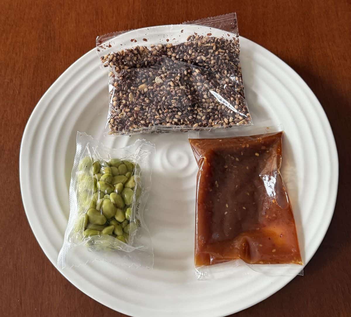 Image showing a packet of crispy rice, a packet of edamame and a pack of salad dressing unopened sitting on a white plate.