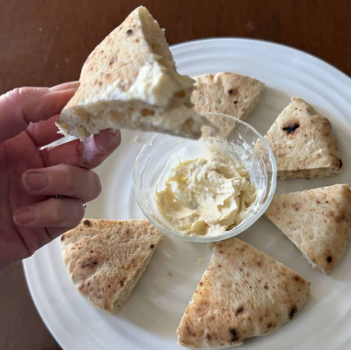 Image of a plate with pita bread on it and a hand holding a piece of pita dipped in hummus over the plate.