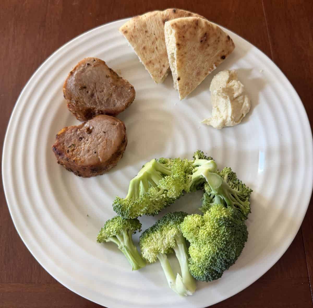 Top down image of the two pork medallions cooked and served on a white plate beside pita bread and hummus and broccoli.