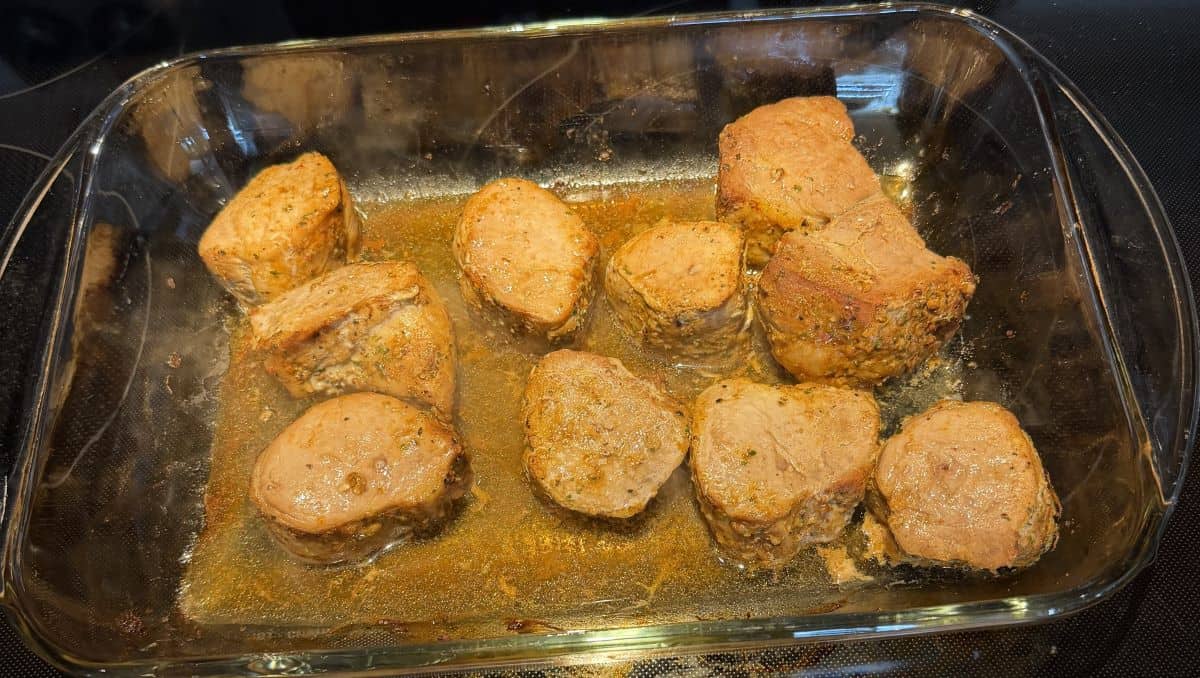 Image of a glass dish with cooked pork tenderloin medallions sitting on top of a stove.