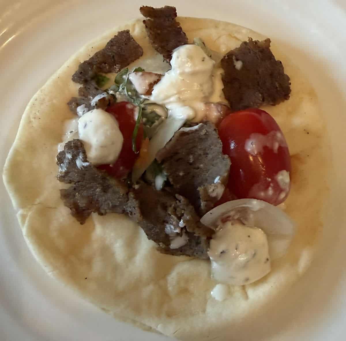 Closeup top down image of one Gyro Pita open faced served on a white plate with tomatoes and tzatziki.