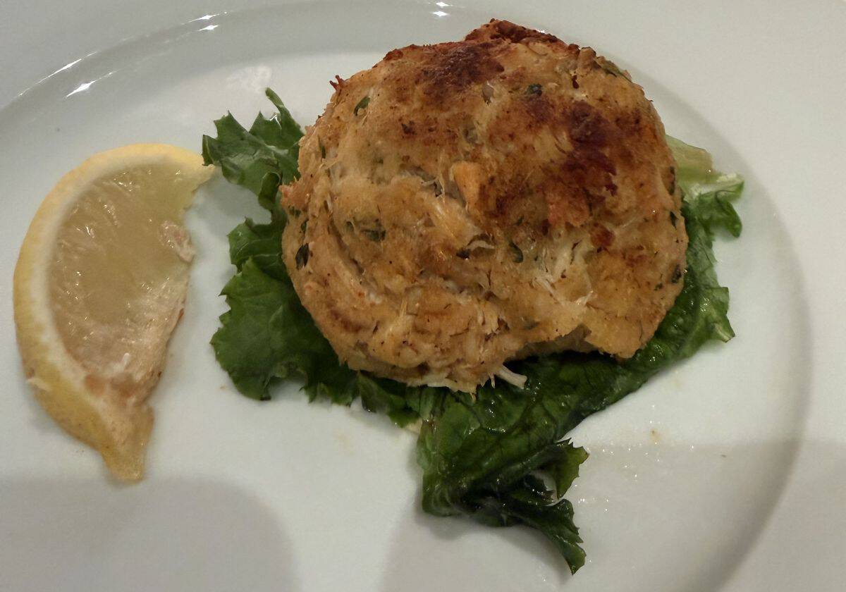Top down image of one crab cake served on a white plate with a leaf of lettuce under it and a lemon wedge beside it.