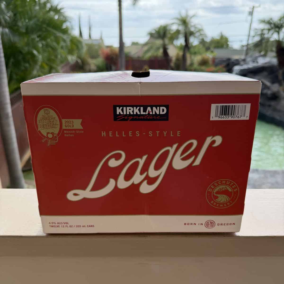 Image of a case of the Kirkland Signature Lager from Costco sitting on a patio unopened. In the background is a pool and palm trees.