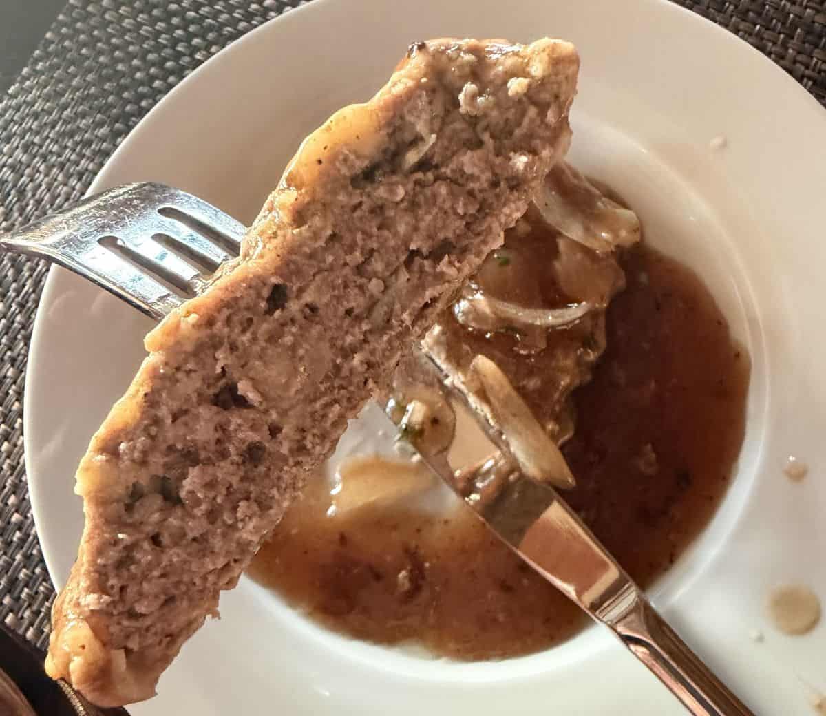 Closeup image of a fork with a piece of burger steak on it hovering over a plate.