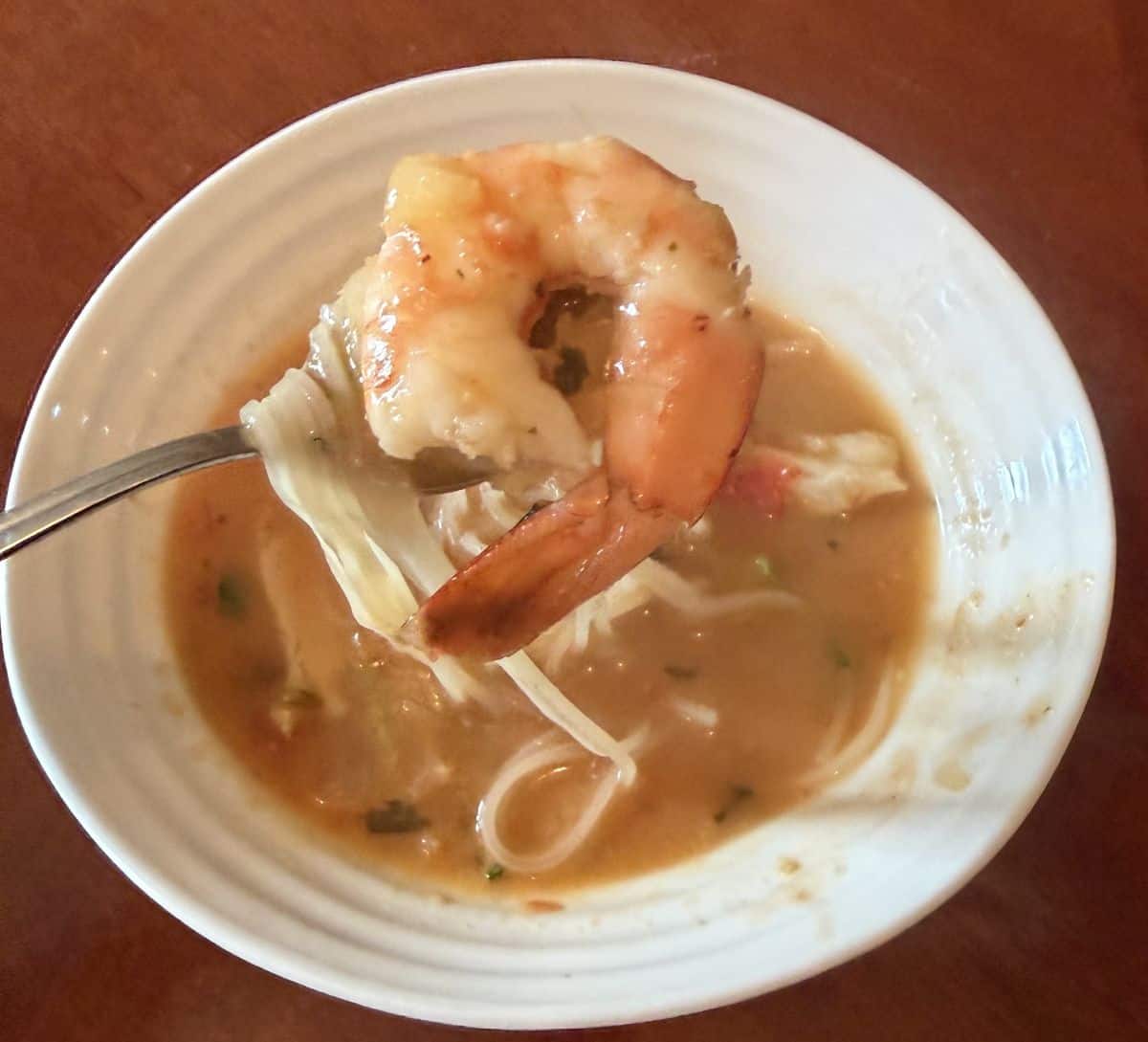 Image of a spoon with shrimp and noodles on it hovering over a bowl of Tom Yum Soup.
