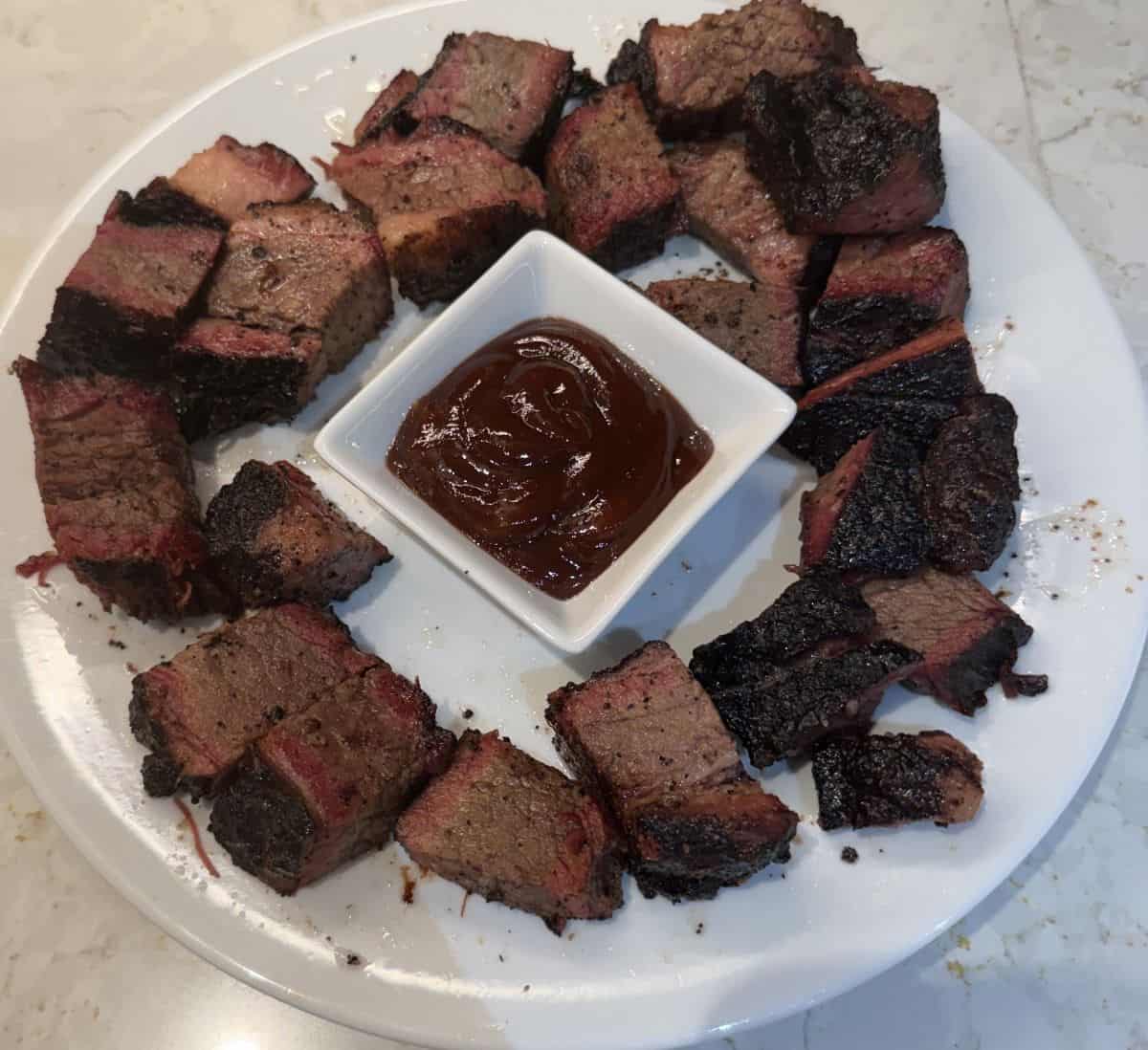 Image of a plate with heated burnt ends on it and in the center of the plate is barbecue sauce for dipping.