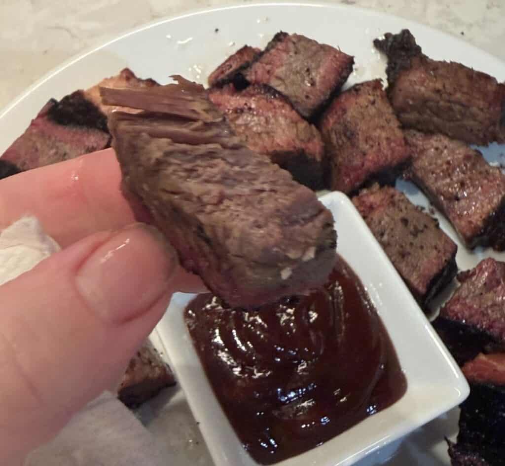Image of a hand holding one burnt end piece close to the camera with a plate of burnt ends in the background.