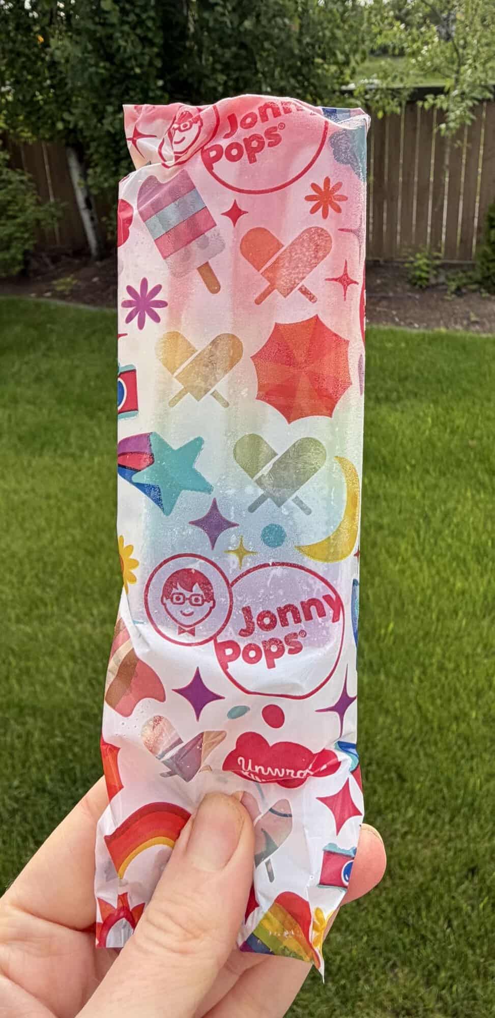 Image of a hand holding an individually wrapped Jonny Pop unopened with grass in the background.