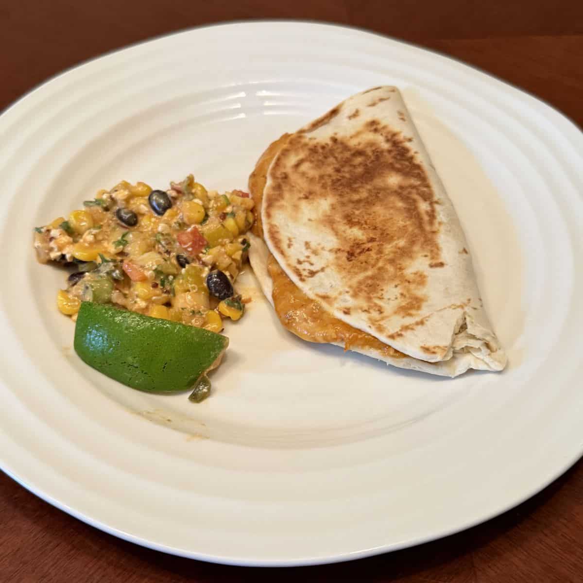 Sideview top down image of a quesadilla beside corn salad and served on a white plate.