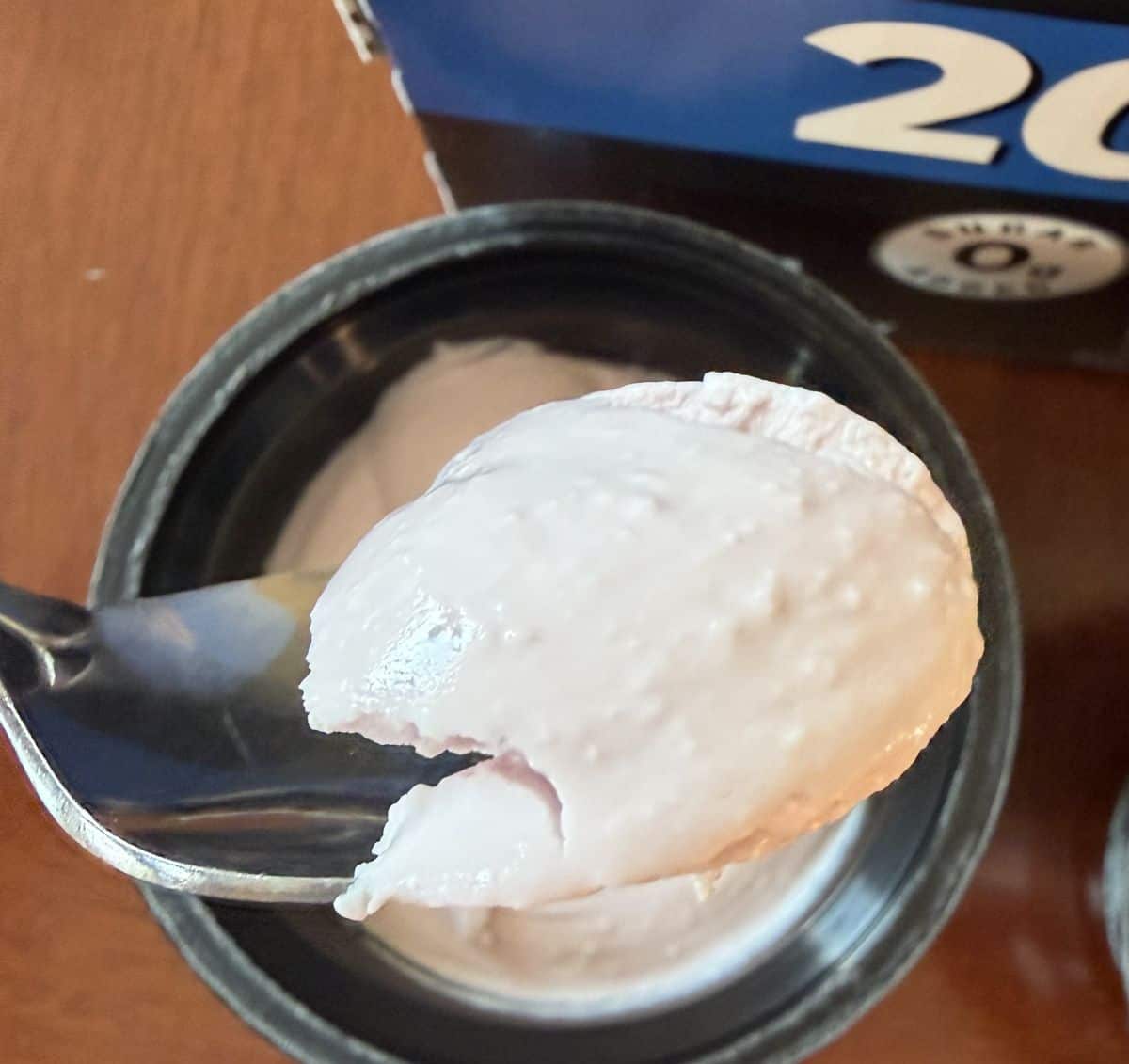 Image of a spoon hovering over a container of yogurt with slightly pink yogurt on the spoon.