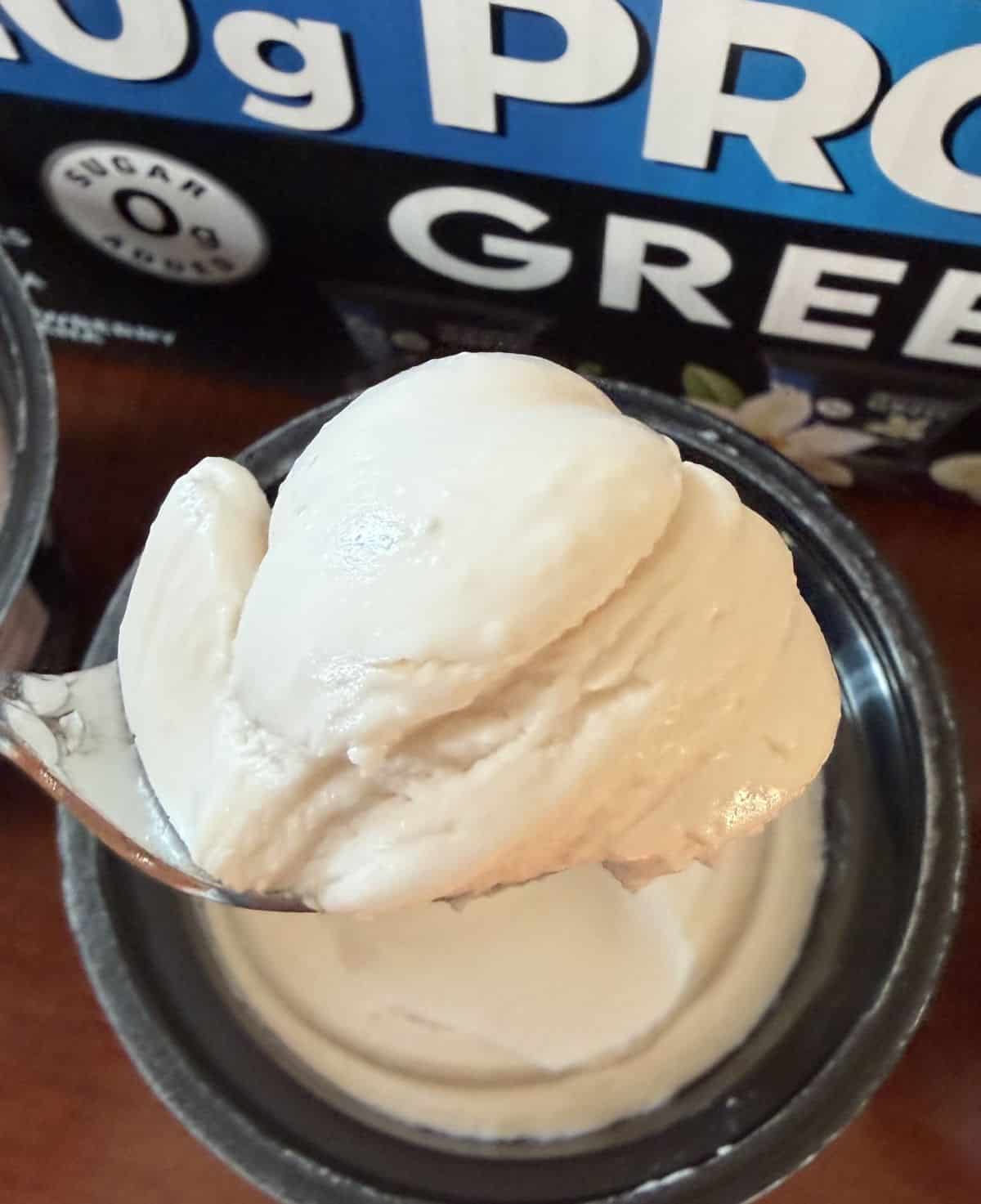 Image of a spoon hovering over a vanilla yogurt container with yogurt on it close to the camera.