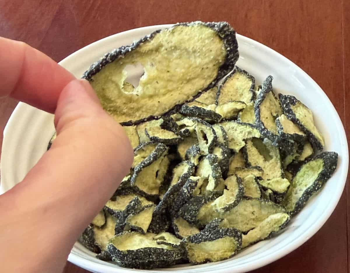 Image of a hand holding one cucumber crisp close to the camera with a bowl of cucumber crisps in the background.