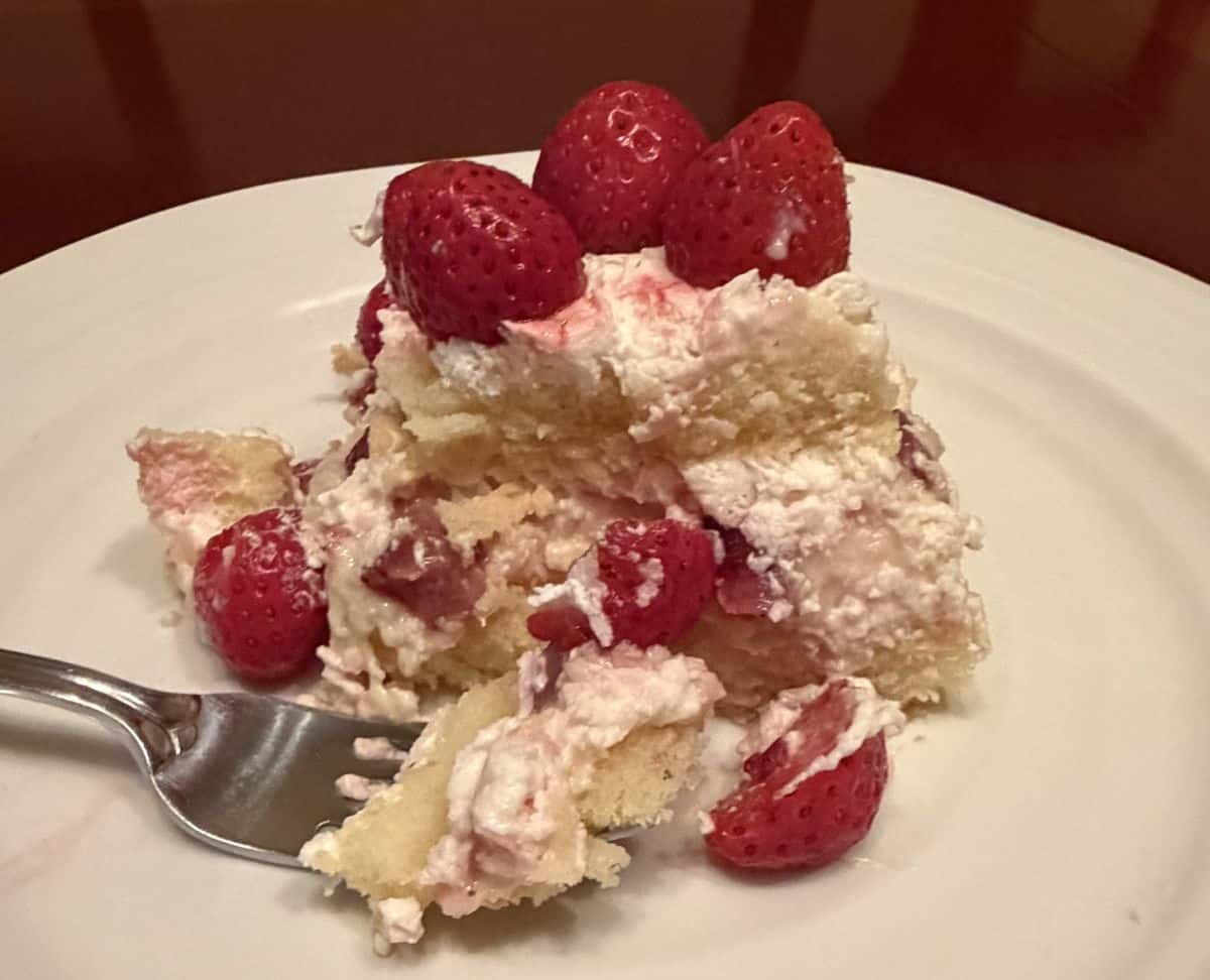 Sideview image of one slice of strawberry scoop cake served on a white plate so you can see the layers in the cake and a fork has a bite of cake on it on the plate.