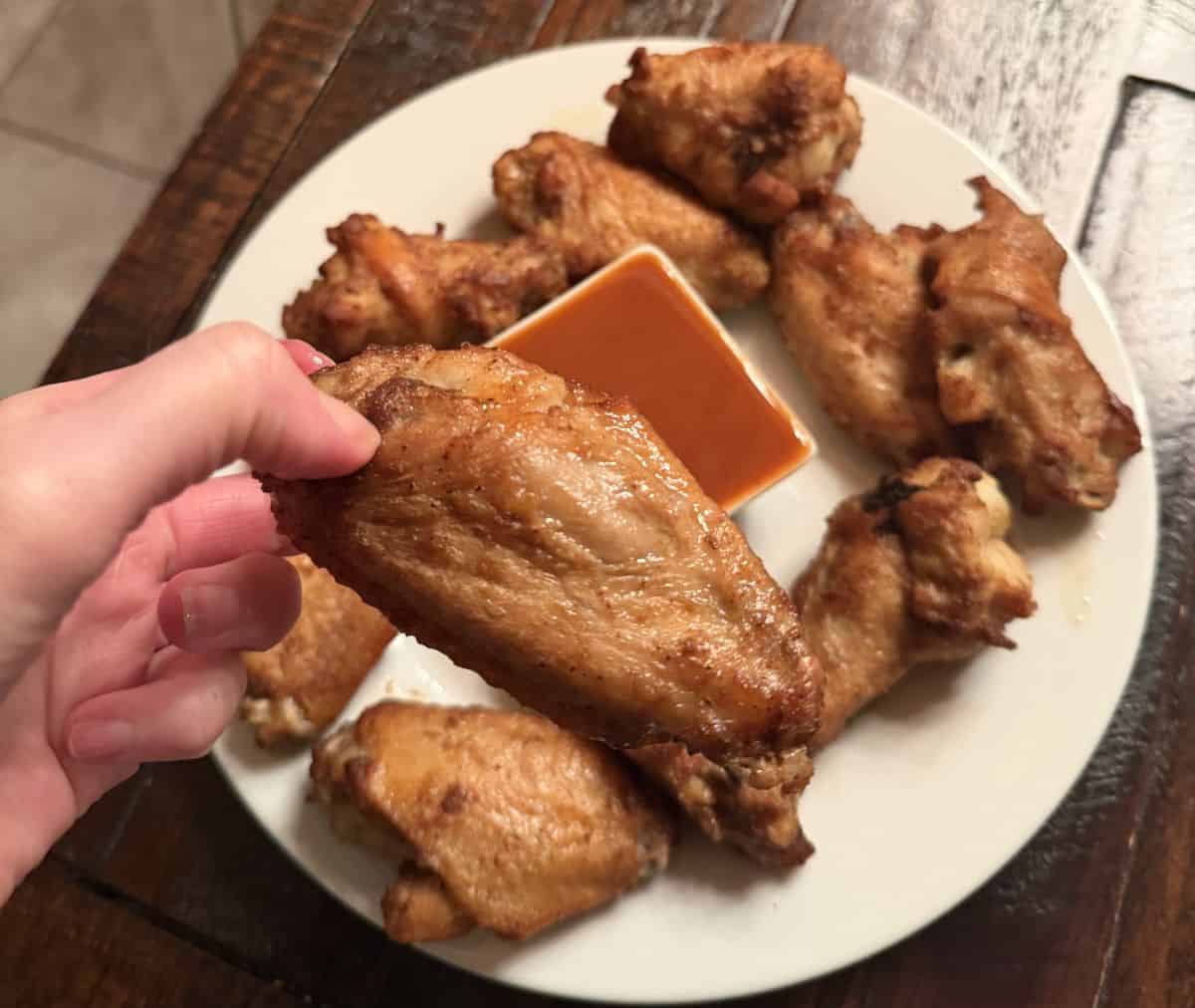 Image of a hand holding one chicken wing close to the camera hovering over a plate of wings.