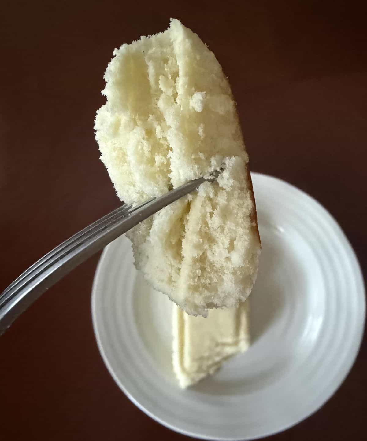 Image of one bite of cheesecake on a fork close to the camera hovering over a plate with a slice of cheesecake on it.