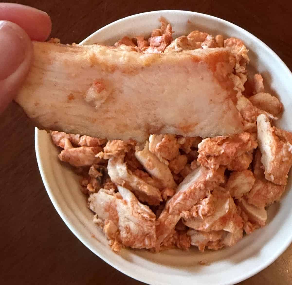 Image of a hand holding one large piece of Kirkland Signature Grilled Chipotle Chicken hovering over a bowl of chipotle chicken.