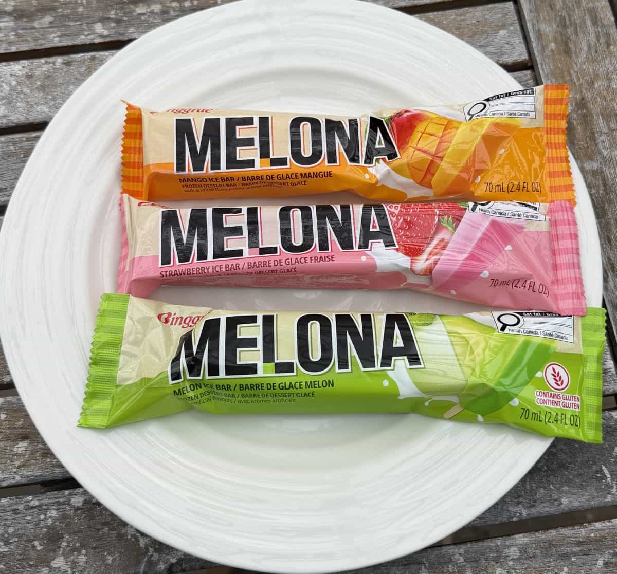 Image of three individually wrapped Melona bars on a white plate, there is strawberry, mango and honeydew.