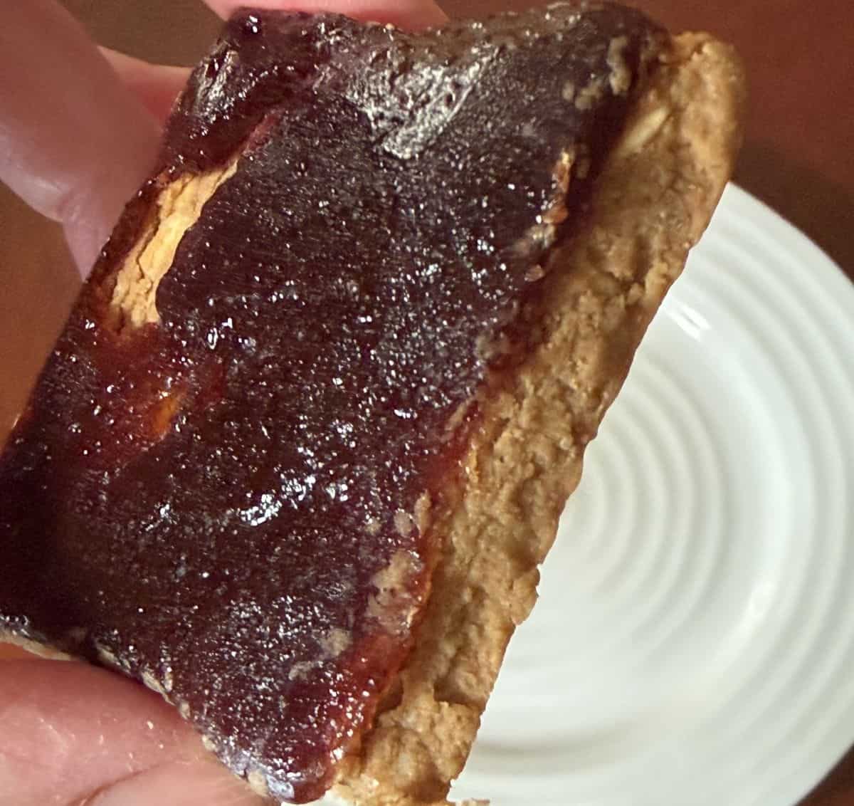 Image of a hand holding one PB & J Mid-Day Square close to the camera so you can see the layer of peanut butter and jelly.