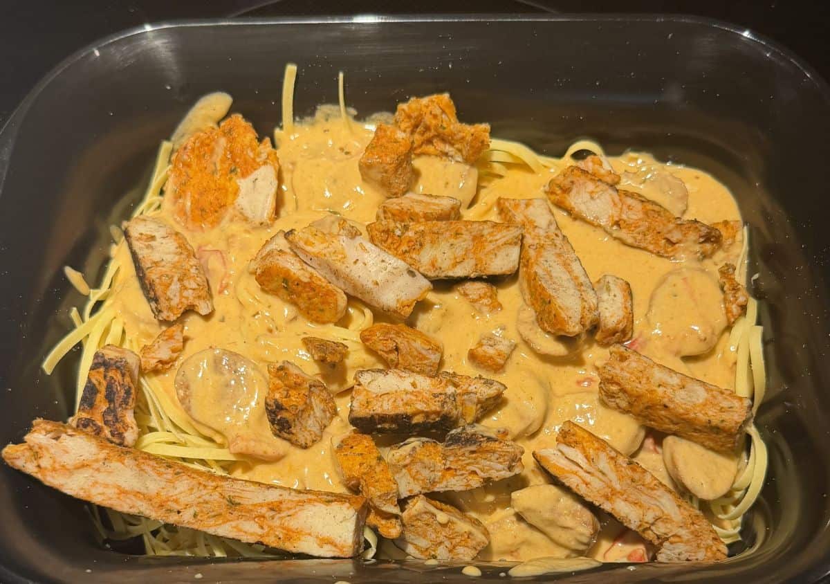 Top down image of a plastic tray sitting on a stovetop with linguini, sauce, sausage and chicken on it before heating it.