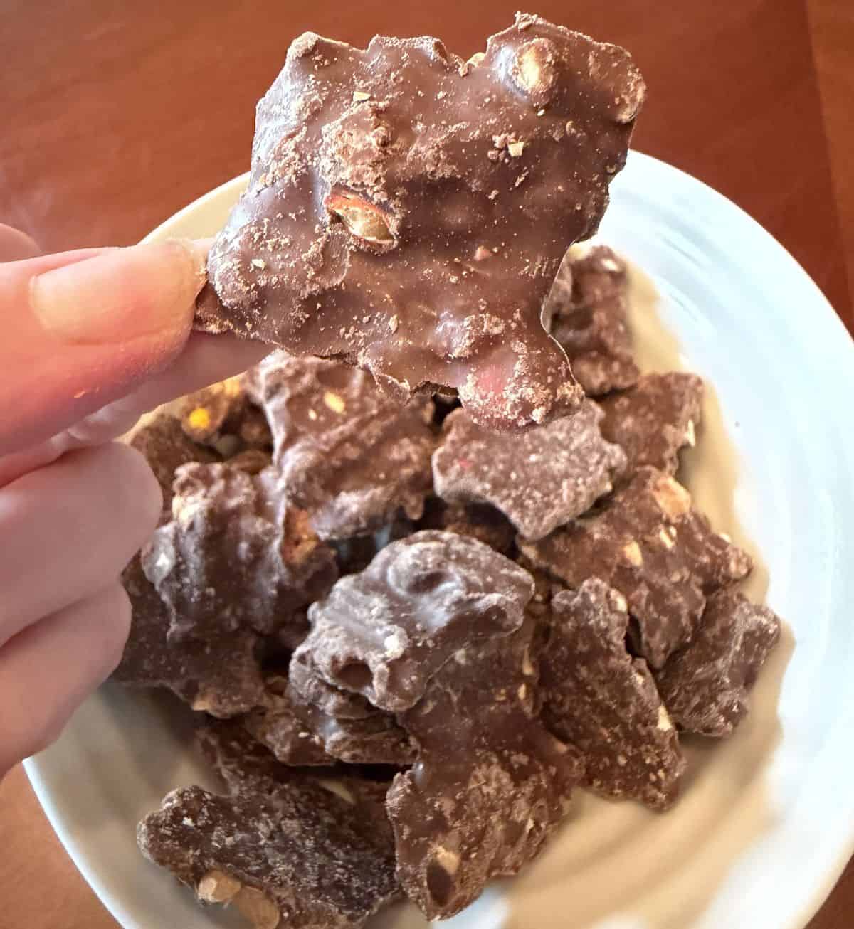 Image of a hand holding one piece of Reese's Pieces Bark hovering over a bowl of bark.