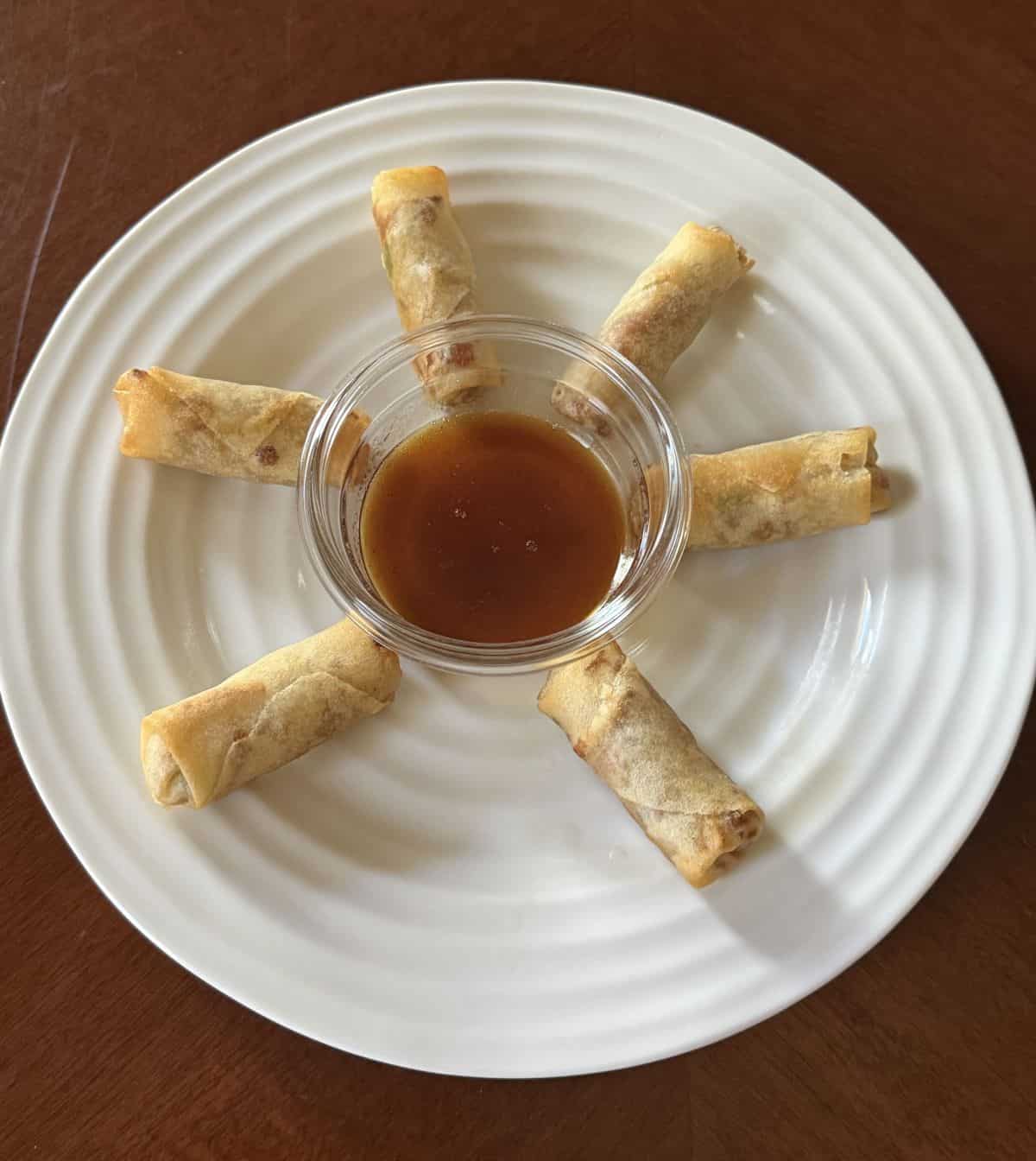 Top down image of six heated spring rolls served on a white plate with a dipping sauce in the middle.