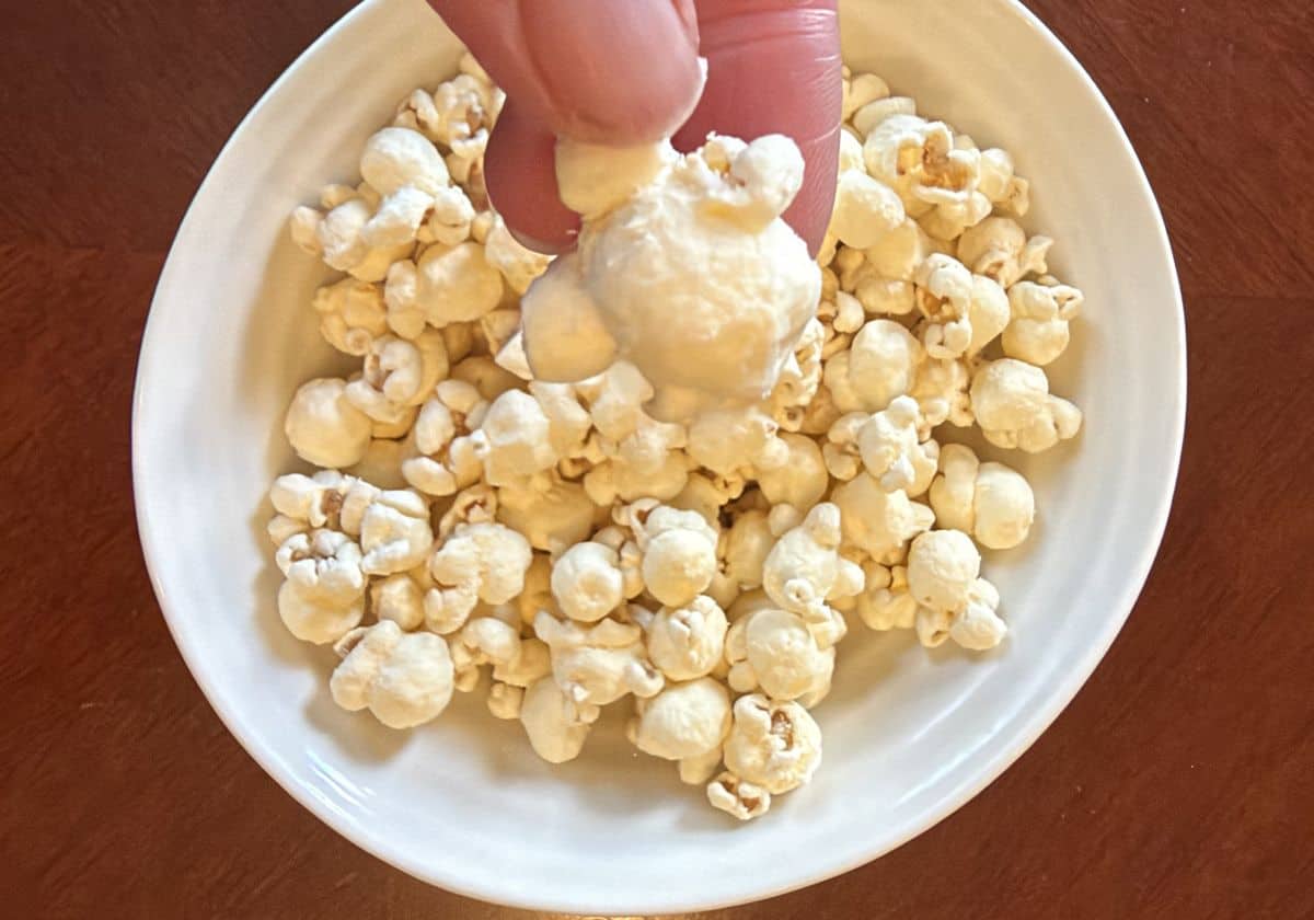 Image of a hand holding one piece of popcorn close to the camera hovering over a bowl of popcorn.