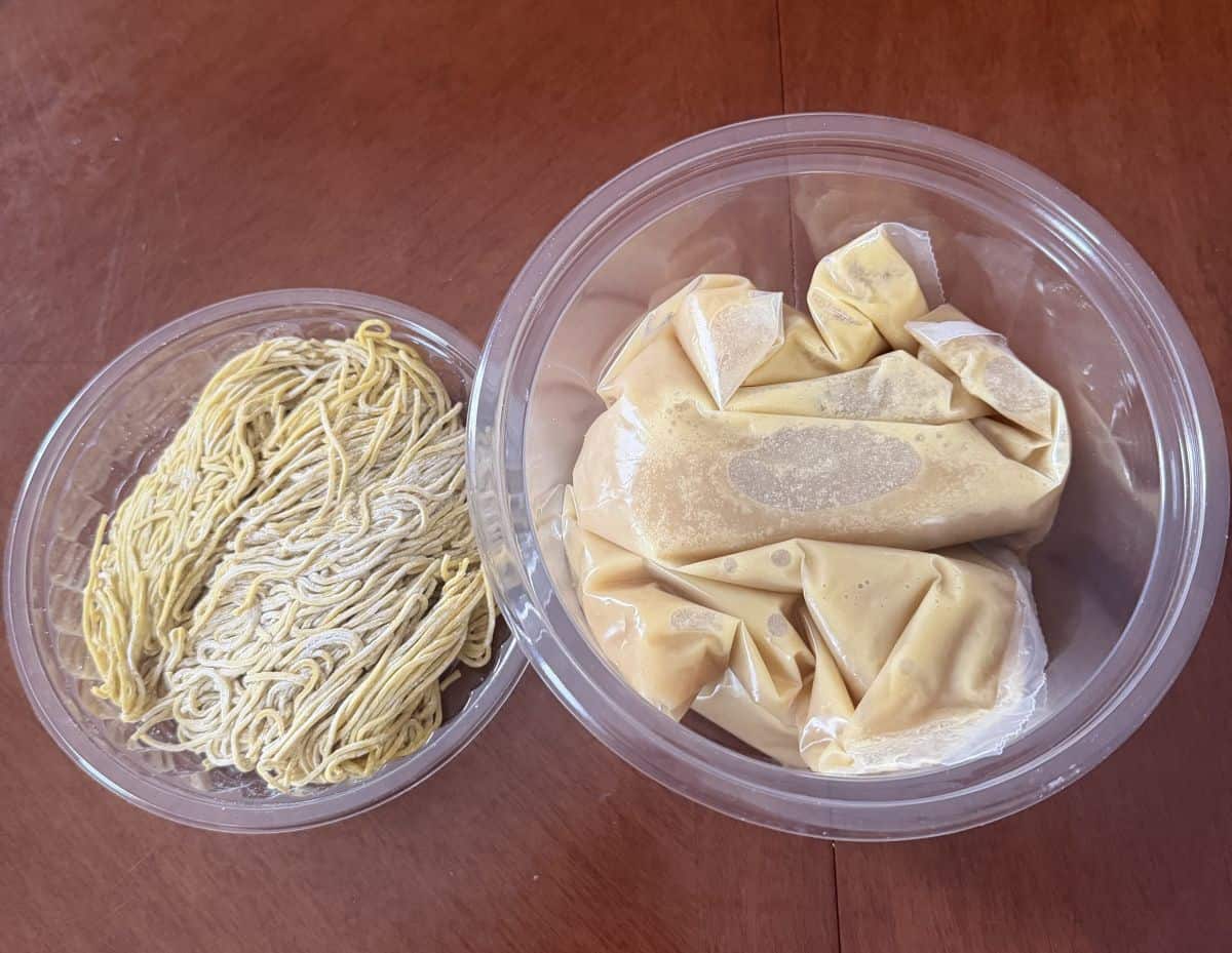 Top down image of the container with the ramen in it showing the broth and noodles. The broth is in a plastic bag and the noodles are in a tray.