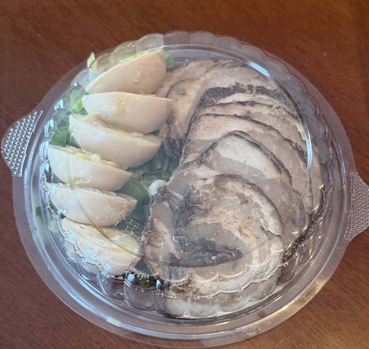 Top down image of the unopened container of ramen showing pork belly, eggs and onions in the top tray.