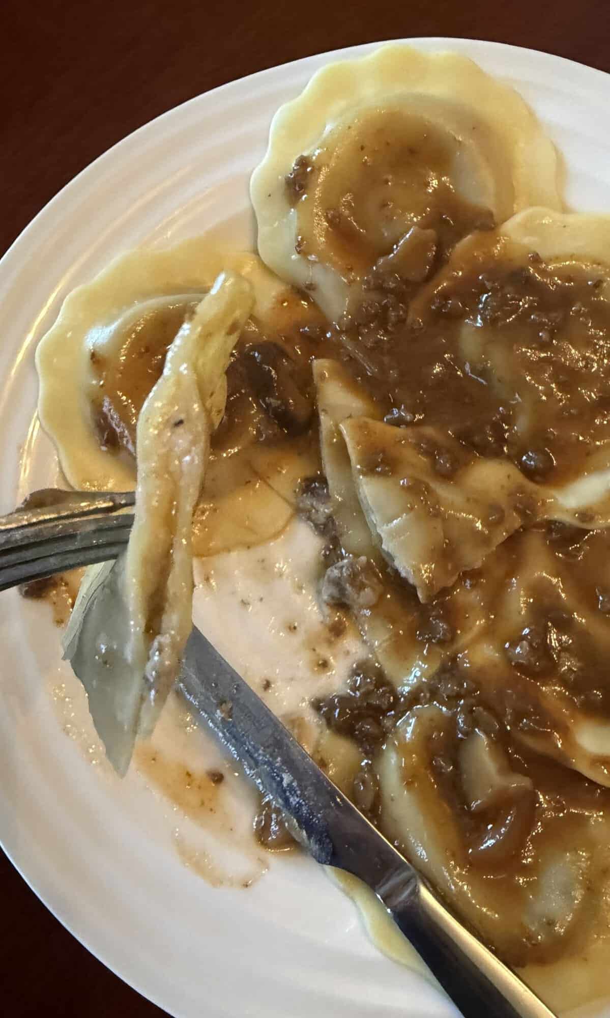 Image of a fork with a ravioli cut in half on it so you can see the filling. Hovering over a plate of ravioli with sauce.